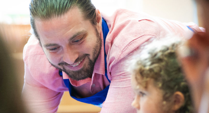 A Crayola Imagine Arts Academy art instructor smiling as he stands next to a boy sitting at his desk