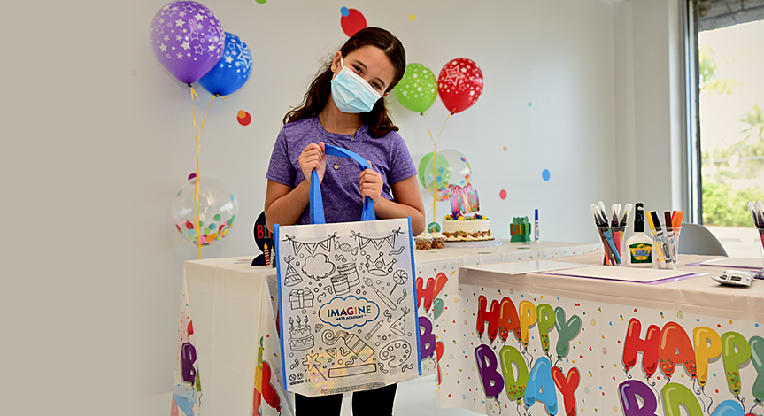 A masked girl at a birthday party holding a canvas color-me tote loot bag.